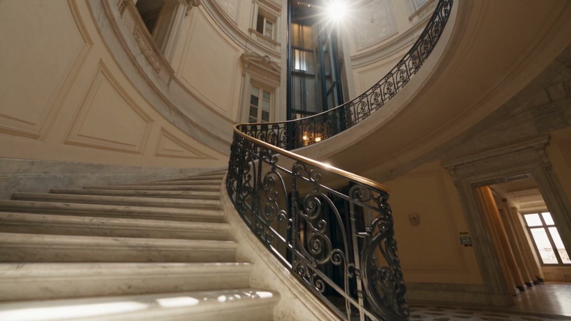 La scala elicoidale di un palazzo storico con l'ascensore di vetro al centro, visto dal basso